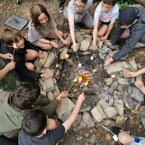 Young children on a day camp surrounding a fire pit burning marshmallows
